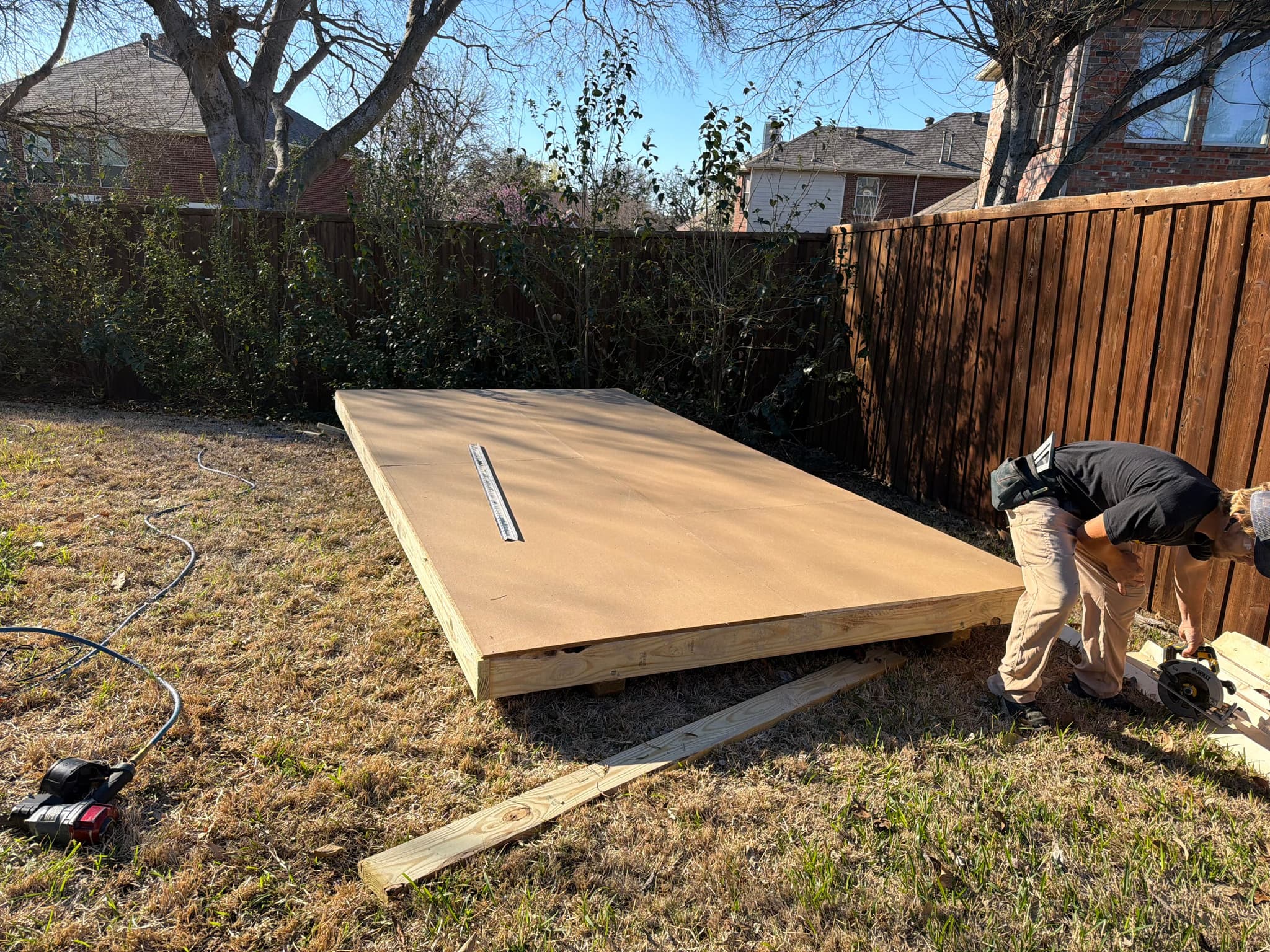 Two builders set an 8x16 wooden floor frame on a Denton backyard lawn beside a wood privacy fence.