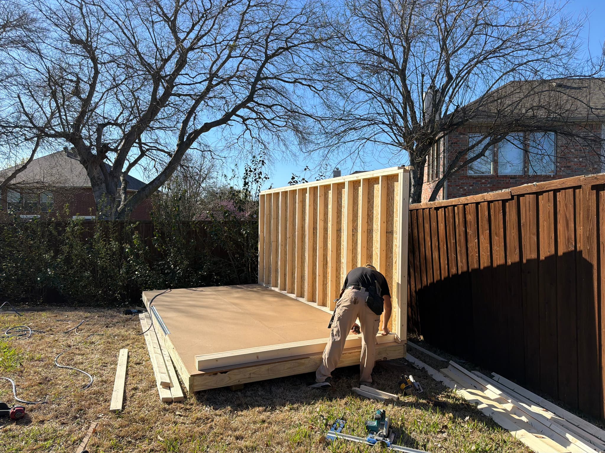 First wall frame raised on a built-on-site shed floor in a fenced Denton backyard.