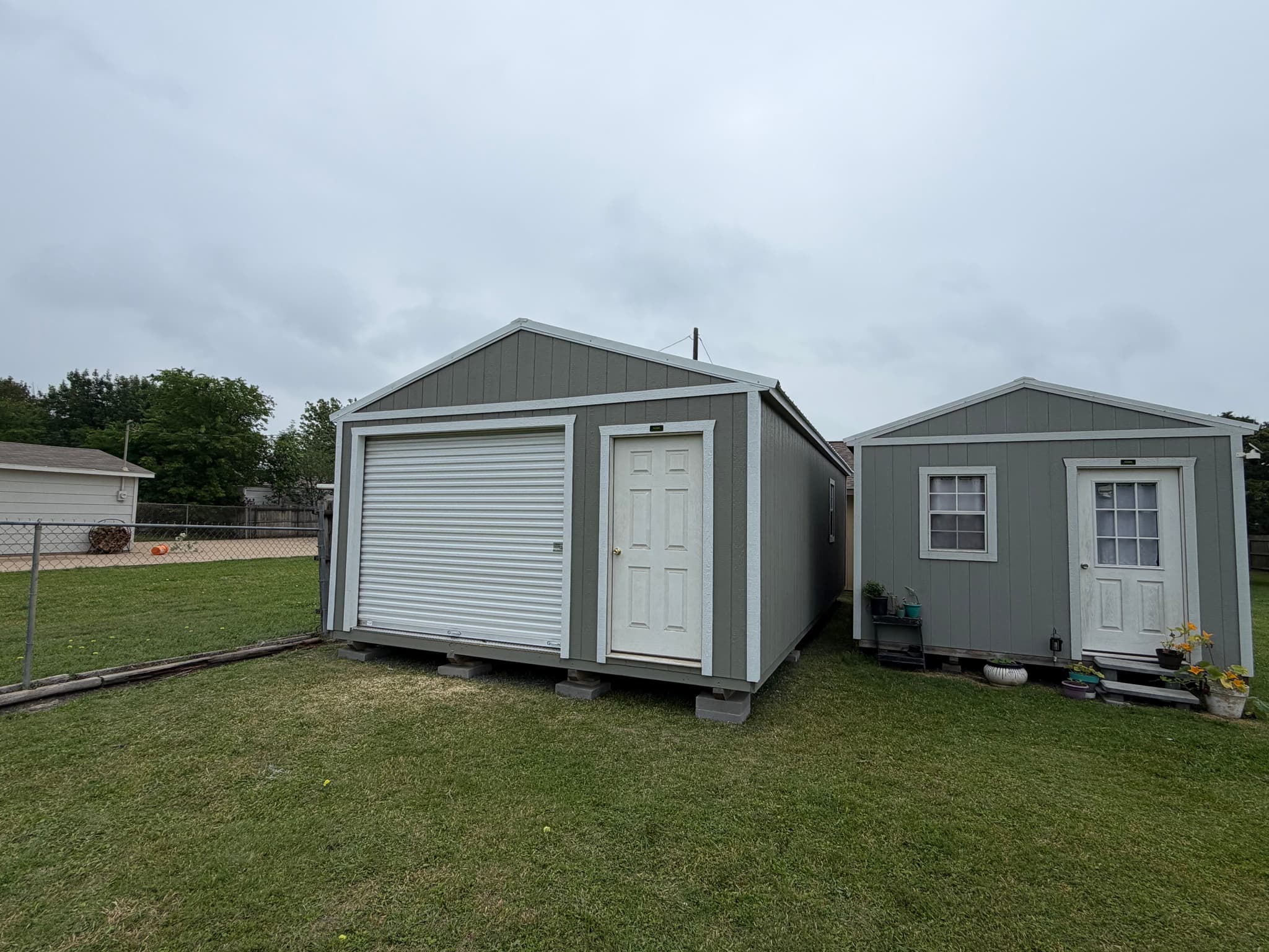 Finished gray utility shed with white roll-up garage door beside a smaller matching gray shed.